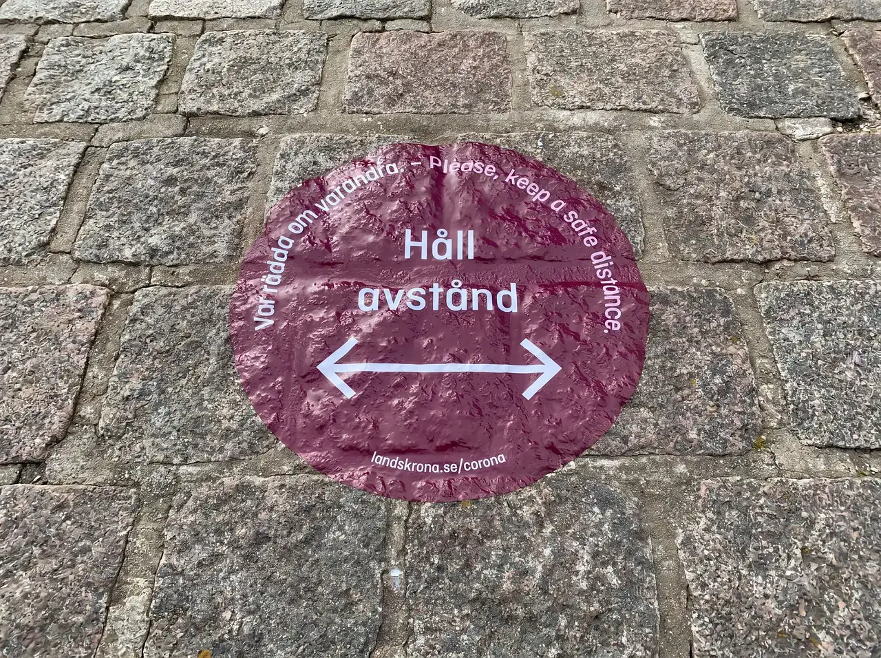 Röd skylt på gatusten med texten "Håll avstånd" och pilar.